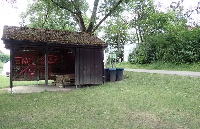 Holzunterstand mit Bank und Graffiti, daneben Mülltonnen und Waldweg im Grünen