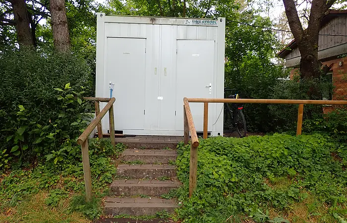 Außenansicht eines weißen Containers mit zwei Türen, beschriftet mit „Herren“ und „Damen“, umgeben von Grün und Bäumen.