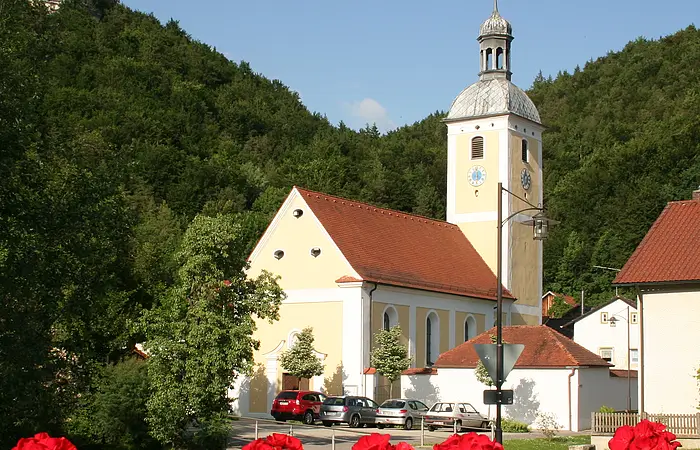 Die Kirche von Mühlbach von der Brücke aus, vorne  Geranien, im Hintergrund der bewaldete Hang.