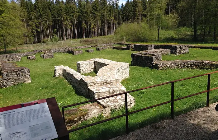 Ruinen von römischen Bauten auf einer grünen Wiese. Im linken unteren Bildrand eine Informationstafel.