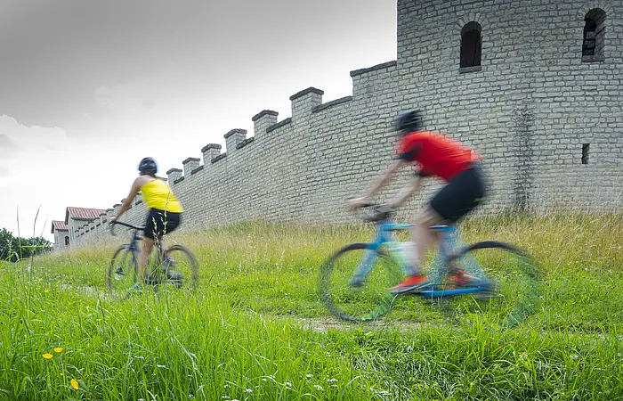 Zwei Gravelbiker vorne mit gelben Shirt, dahinter mit rotem Shirt fahren seitlich am Kastell entlang. Der Himmel ist grau bedeckt, die Wiesen saftig grün.