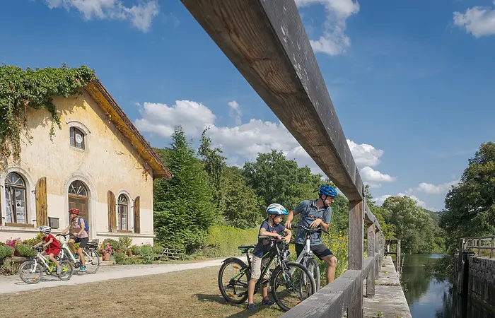 Familie mit vier Personen und Fahrrädern neben einem alten Haus und einem Fluss unter blauem Himmel.