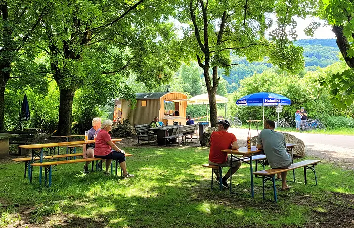 Menschen sitzen an Holzbänken unter Bäumen in einem schattigen Biergarten mit Imbisswagen im Hintergrund.