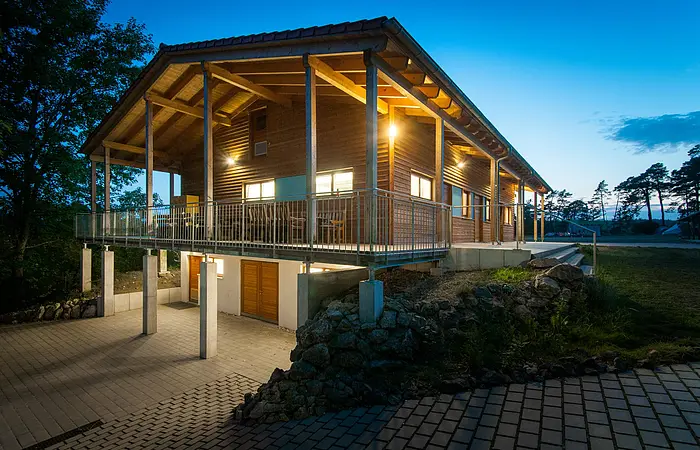 Ein Holzhaus mit großer Terrasse ringsherum ist hellbeleuchtet in der Nacht. Ein gepflasteter Weg führt links hinunter zum selben Gebäude, das im Untergeschoss gemauert und im Hang gebaut ist. Die Terrasse wird durch Pfeiler gestützt.