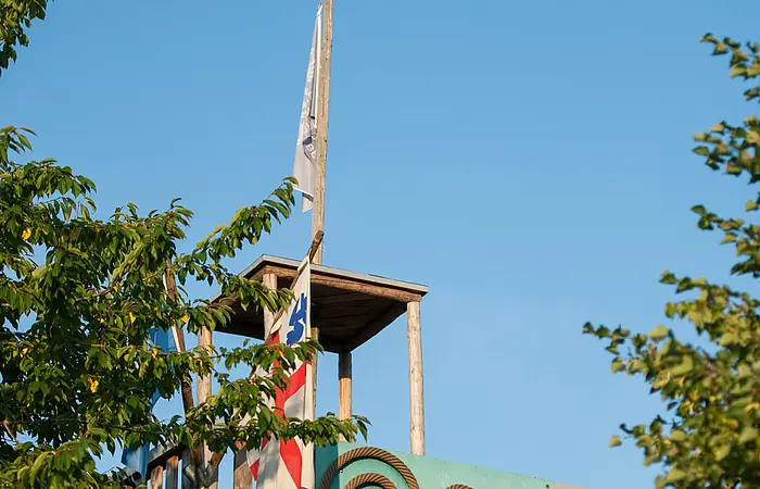 Eingerahmt von zwei großen Bäumen steht ein Aussichtsturm mit einem Kunstobjekt auf grünem Hintergrund. Links ist eine Fahne gehisst.