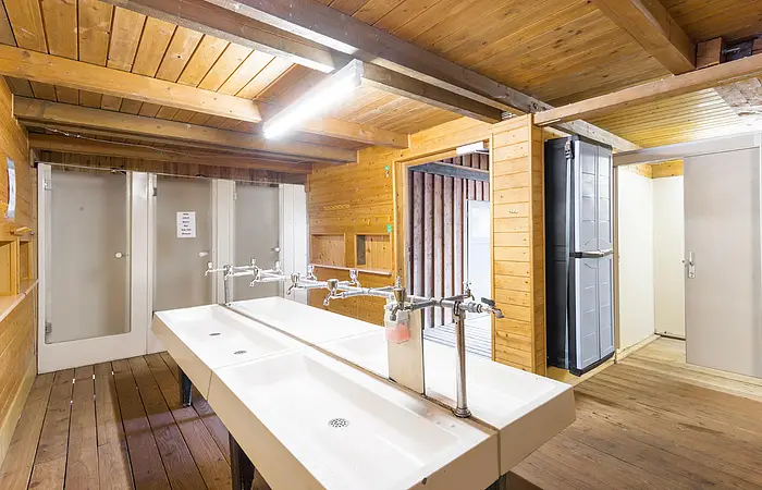 Der Waschraum hat zwei große Waschbecken in der Mitte . Rechts geht es zu den Duschen und WC's. Das Gebäude ist aus Holz gebaut.