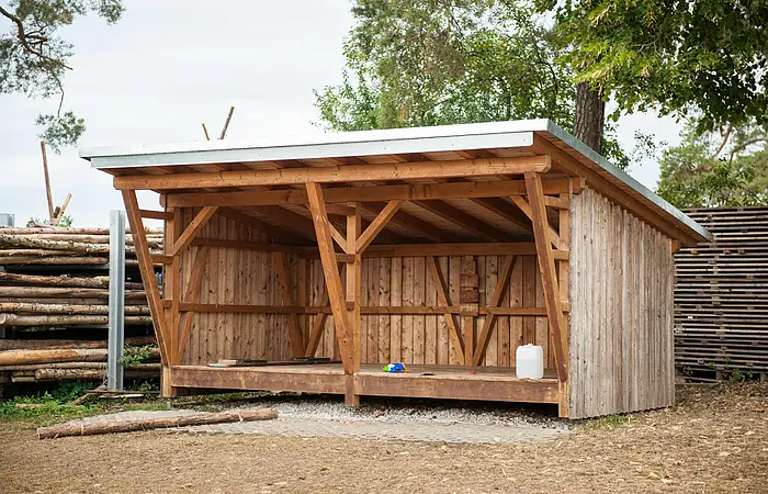 Eine Schutzhütte aus Holz steht vor einem Holzzaun. Dahinter stehen einige Bäume.