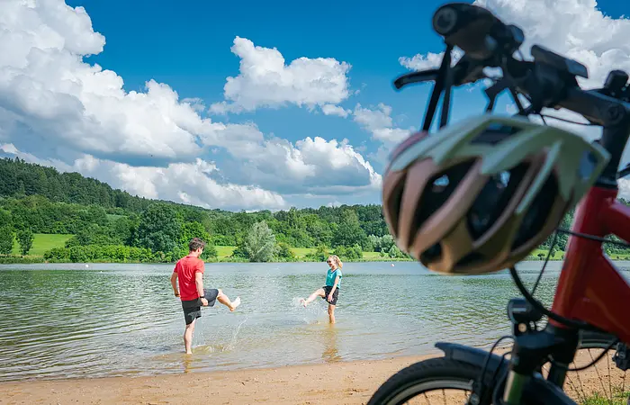 Im Vordergrund stehen rechts zwei Fahrräder, dahinter liegt ein kurzer Strand an einem Seeufer. Im seichten Wasser stehen ein Mann und in eine Frau in kurzen Hosen und T-Shirts, die versuchen, sich gegenseitig nass zu spritzen. Am Ufer gegenüber liegen eine Wiese und Wald.