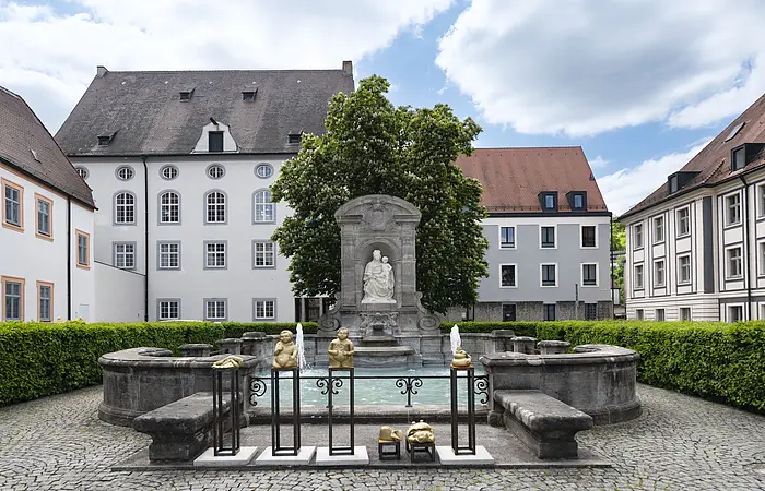 Zentrum des Bildes ist eine Ädikula mit einer Steinstatue, die die heilige Maria, die Patrona Bavaria, mit Kind darstellt. Unmittelbar davor befindet sich das rechteckige Brunnenbecken, welches von einer kniehohen Stein-Mauer begrenzt wird.
Dahinter stehen ein großer Baum und niedrige Hecken, die den Kiesplatz, auf dem der Brunnen steht, einsäumen.
Im Hintergrund befinden sich verschiedene mehrstöckige Häuser in hellen Farben.