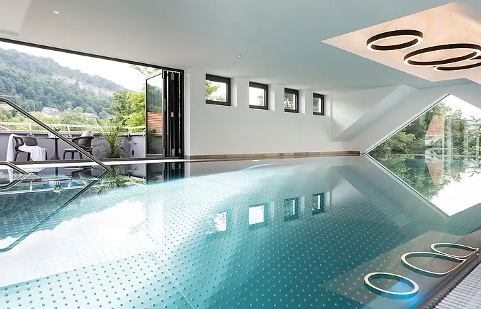 Innenraum eines modernen Hallenbads mit großen Fenstern und Blick auf eine Terrasse und bewaldete Hügel.