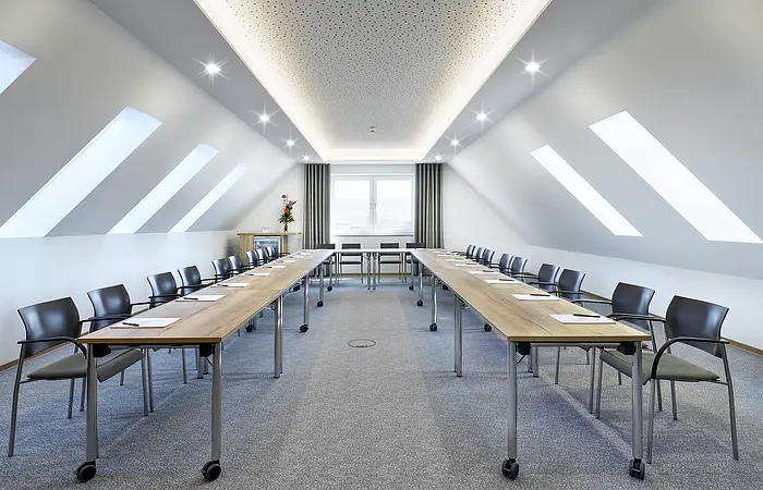 Konferenzraum mit zwei langen Tischen, Stühlen und Dachfenstern in einem hellen Raum mit Teppichboden.