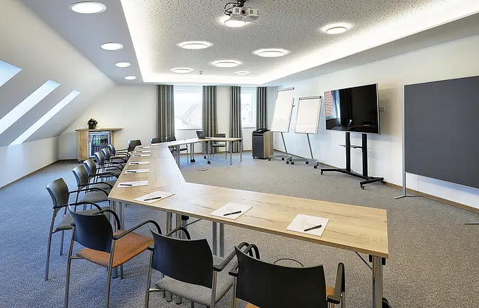 Konferenzraum mit U-förmigen Tischen, Stühlen, Flipcharts, großem Bildschirm und Dachfenstern.