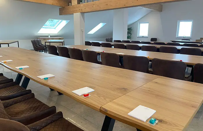 Konferenzraum mit langen Holztischen, braunen Stühlen, Notizblöcken und kleinen Gegenständen auf den Tischen.