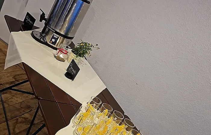 Tisch mit Gläsern und Orangenscheiben, Thermoskanne, Blumen und Kerze vor weißer Wand in Raum mit Holzboden.