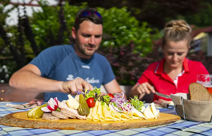 Zwei Gäste genießen üppige Brotzeitplatte im Gasthof "Zum blauen Hecht" in Kipfenberg. Die Platte ist bestückt mit verschiedenen Käse- und Wurstaufschnitten, Radieschen, Tomaten, Essigkuren und roten Zwiebeln. Daneben steht ein Brotkorb.