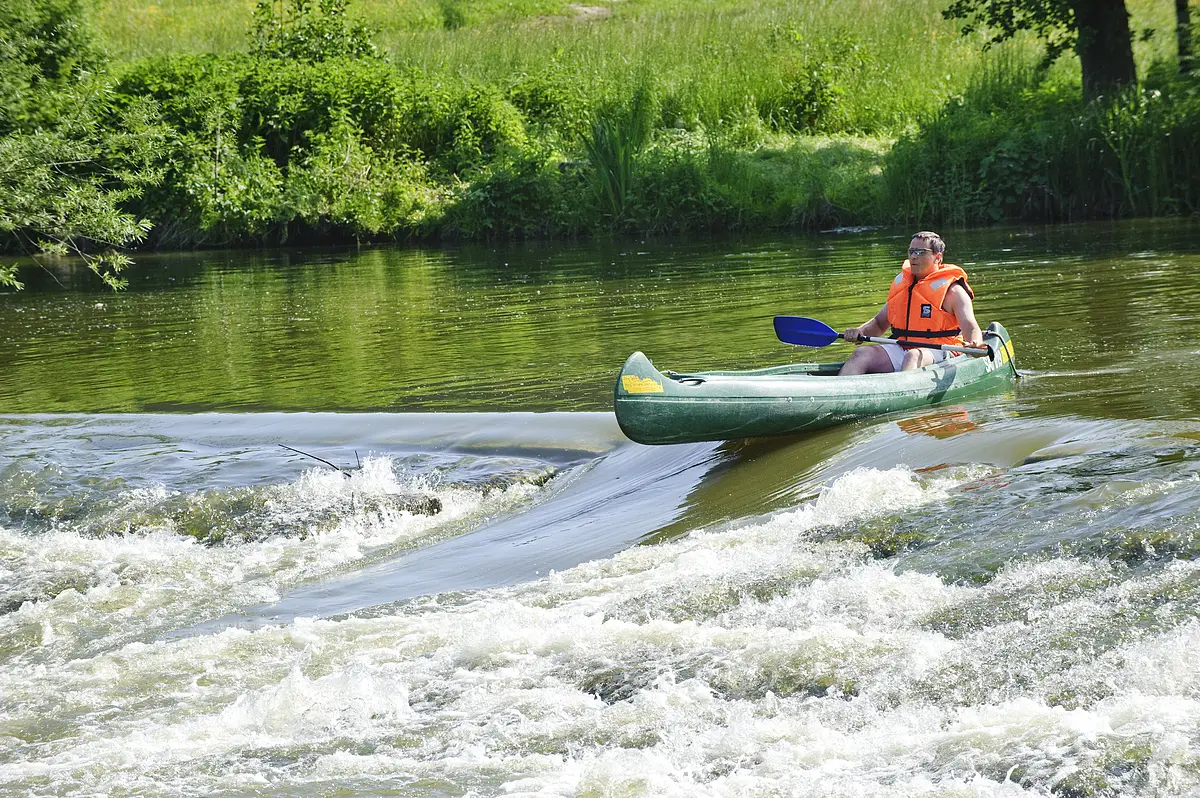 Bootwanderer auf der Bootsrutsche Hammermühle Ein Bootsfahrer mit oranger Rettungweste und Paddel steht mit seinem grünen Kanu am Anfang der Bootstreppe in der Altmühl. Das Wasser rauscht hier an dem steinernen Wehr hinunter. Die Bootsrutsche ist eine Vertiefung für die Fische und auch Boote. Das Ufer ist sattgrün auf dem die Sonne scheint.