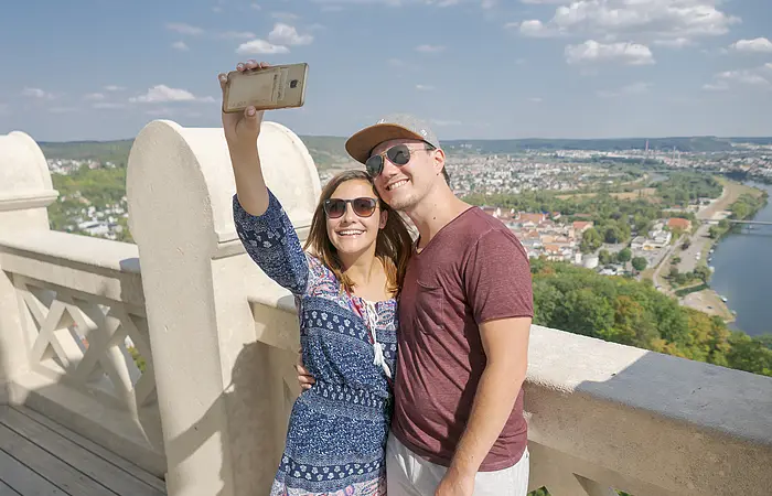 Ein Pärchen steht auf dem oberen Umgang, der um die Befreiungshalle führt. Die Frau hält ihr Smartphone in die Höhe und macht ein Selfie von sich und ihrem Freund, im Hintergrund Kelheim und der blaue, leicht bewölkte Himmel.
