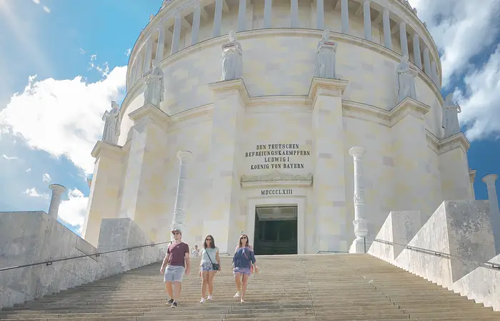Zwei Frauen und ein Mann, in sommerlicher Kleidung, steigen die Treppen der Befreiungshalle hinunter. Der imposante Rundbau mit dessen Eingang ragt hinter ihnen auf. Der Himmel strahlt blau und ein paar Wolken sind zu sehen.