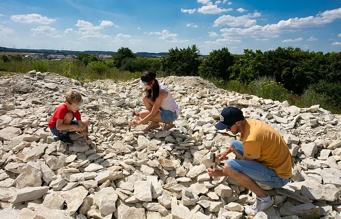 Eine Familie sucht an der Fossiliensammelstelle nach Versteinerungen. Alle drei knien mit Hammer und Meißel und versuchen die Platten zu teilen.