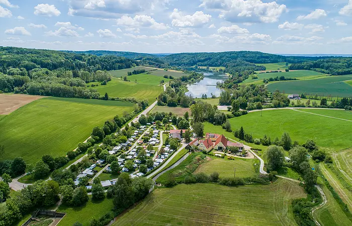 Der Campingplatz Hasenmühle von oben inmitten grüner Wiesen und Bäumen. Hinter dem Platz ist der Hahnenkammsee zu sehen.