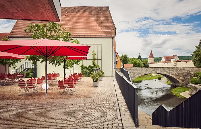 Links unter einem roten Schirm stehen rote Stühle und Tische. Ein grüner Baum lockert alles auf. Rechts fließt die Sulz. Im Hintergrund sind Häuser und die Brücke über die Sulz zu sehen.
