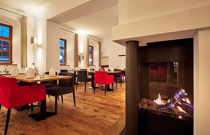 Rote Sessel stehen an Holztischen dahinter sind Fenster. Rechts ist ein Kachelofen mit offenem Feuer, das gemütlich vor sich hin brennt.