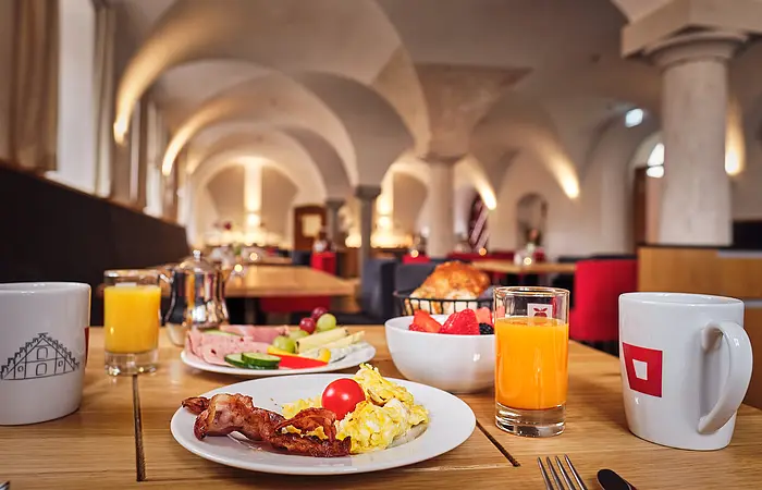 Der Frühstückstisch ist reichlich gefüllt mit Rührei, Speckstreifen, einem Teller mit Wurst und Käse. Rechts ein Glas mit Orangensaft, links steht eines mit Multivitaminsaft. Daneben eine Tasse Kaffee. Schaut man weiter in den Raum erkennt man die beleuchteten Rundbögen des Raumes.