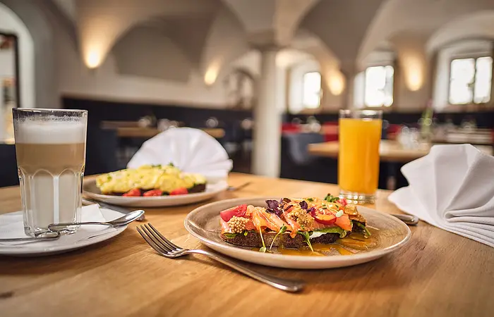 Links steht ein Latte Macciatto dahinter ein Teller mit Rührei. Rechts ein Glas mit Orangensaft gefüllt und eine weiße Serviette zum Fächer gefalten. Im Mittelpunkt ist ein Teller mit belegtem Brot.