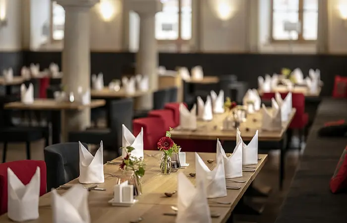 Das Restaurant ist festlich gedeckt mit kleinen Blumenvasen, weißen Servietten. Die dunklen Stühle haben rote Polster. Der Raum hat zwei weiße Säulen und viele Fenster.
