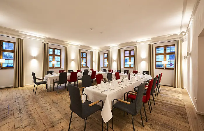 Eine große festlich gedeckte Tafel in U-Form steht in diesem hellen Raum mit vielen Fenstern. Die Tafel ist mit weißen Tischdecken und Servietten eingedeckt.
