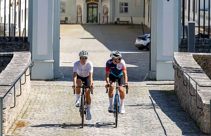 Zwei Rennradler fahren durch das Tor von Schloss Hirschberg bei Beilngries, in deren Hintergrund erkennt man den zentralen Eingang des Schlosses