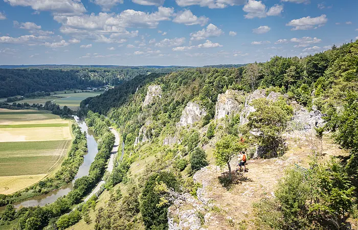 Sommerlicher Blick ins Altmühltal auf das Tal, auf den Fluss und auf die Felsenkette Arnsberger Leite, auf der ganz klein am Talrand eine Familile steht und ins Tal schaut.