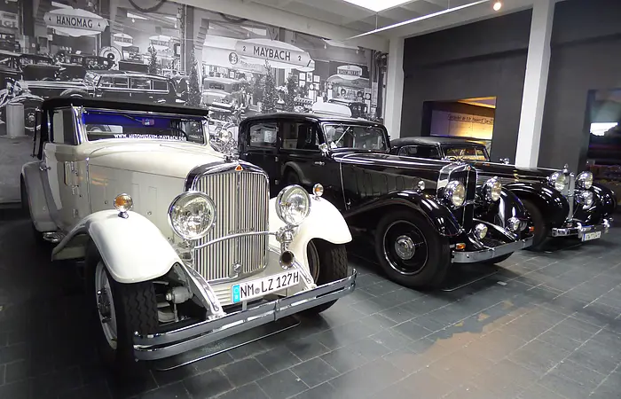 Drei Oldtimer-Autos nebeneinander in einem Museum mit historischem Schwarz-Weiß-Foto im Hintergrund.