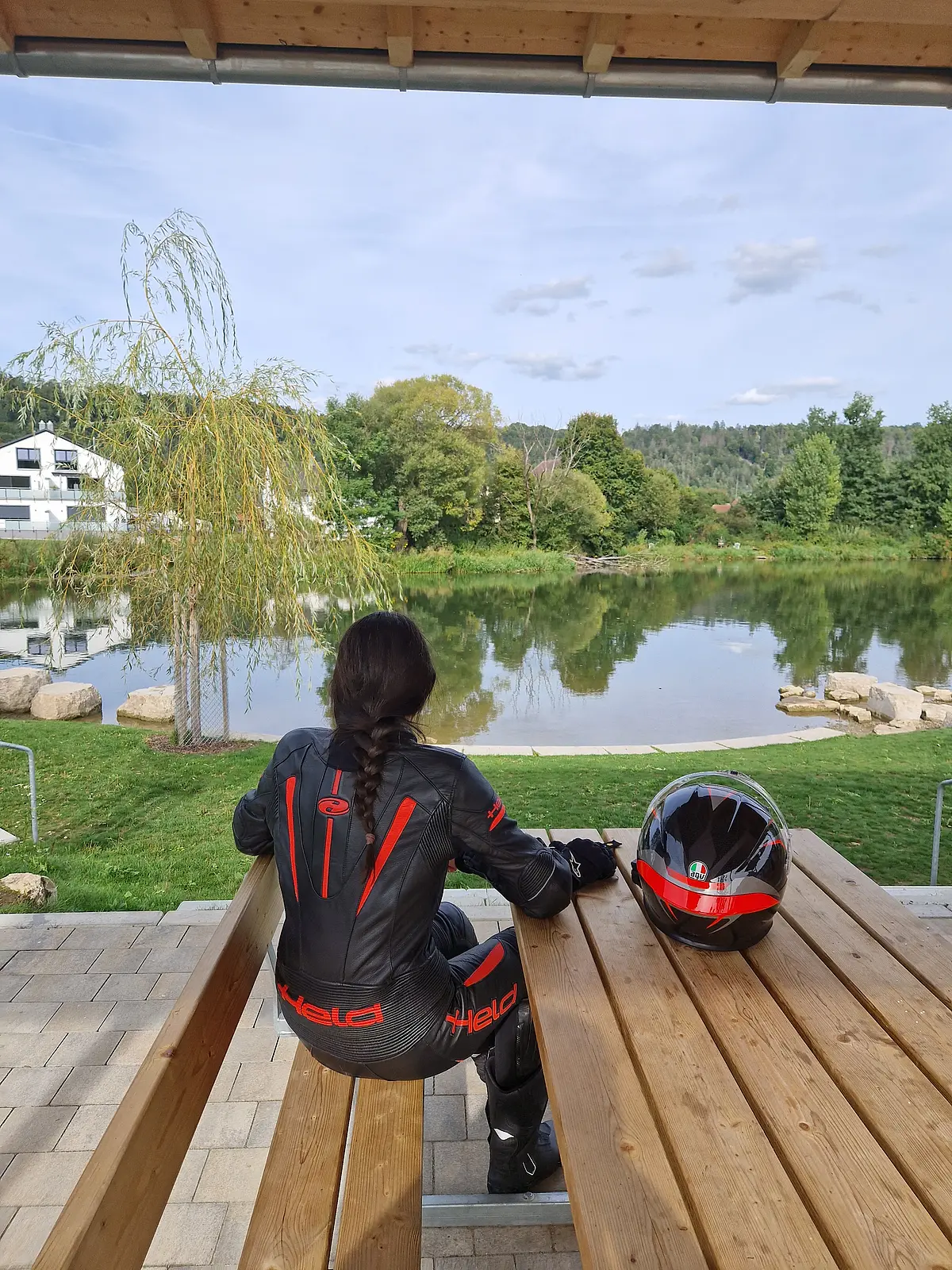 Motorradfahrerin in Kottingwörth Person in schwarzer Motorradkleidung sitzt auf Holzbänken unter einem Dach und blickt auf einen See mit Bäumen im Hintergrund.