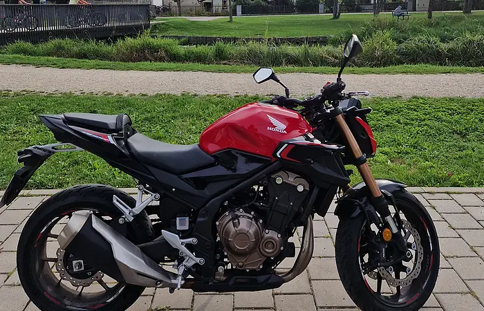 Motorrad der Marke Honda in rot und schwarz auf gepflastertem Weg vor Park mit Bäumen und Häusern im Hintergrund.