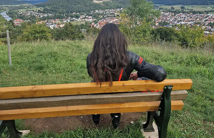 Person mit langen dunklen Haaren sitzt auf einer Bank und blickt auf eine Stadt im Tal unter bewölktem Himmel.