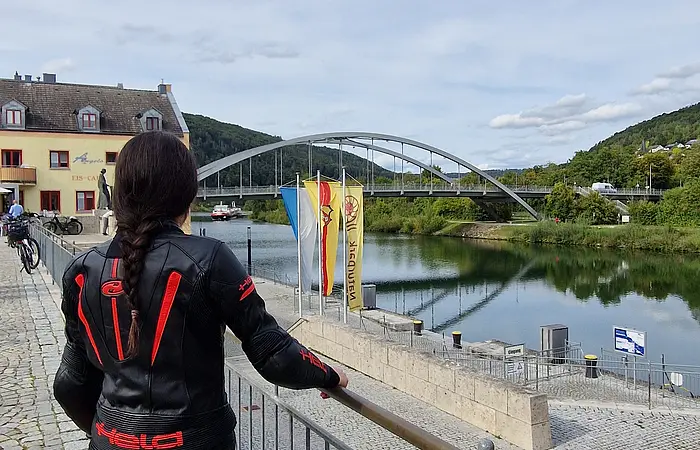 Person in schwarzer Motorradkleidung mit rotem Helm blickt auf Fluss, Brücke und Gebäude bei bewölktem Himmel.