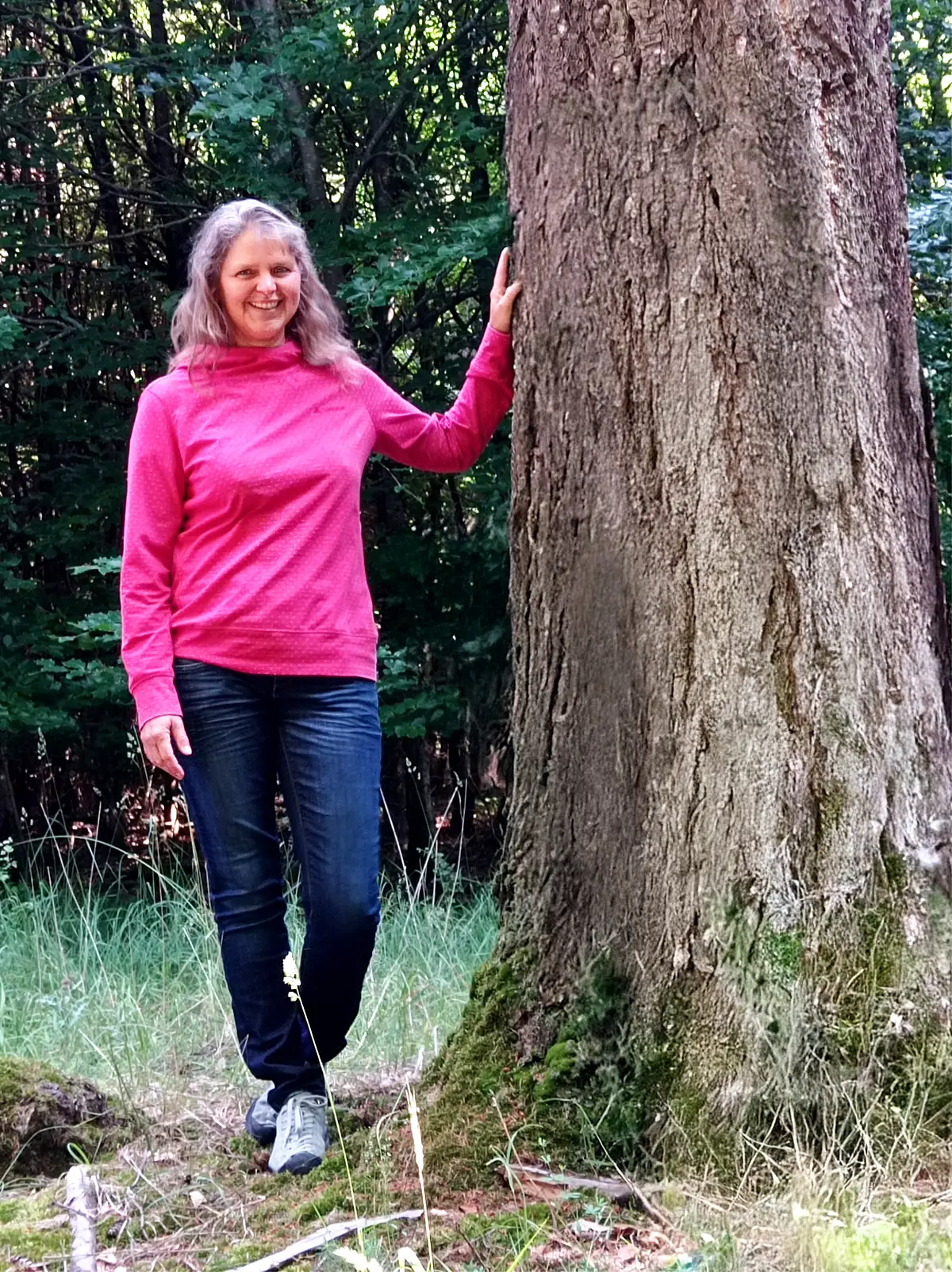 Eine Frau mittleren Alters mit langen, lockigen, blonden Haaren steht im Wald neben einem dicken Baum. Sie hat eine Hand an den Stamm gelegt. Sie trägt ein pinkfarbenes Langarmshirt, eine dunkelblaue Jeans und graue Schuhe. Die Frau lächelt freundlich in die Kamera. Der Hintergrund besteht aus dichtem, grünem Laub, das von Sonnenlicht durchbrochen wird.