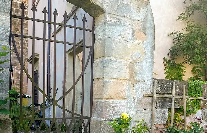 Ein Tor aus Metall in einem Gemauerten Steinbogen. An die Mauer schließt ein Haus an.
