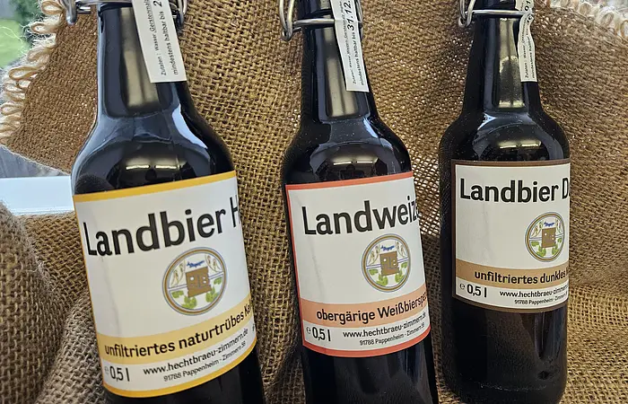 Drei braune Bierflaschen mit Bügelverschluss und Etiketten vor grobem Jutestoff stehend.