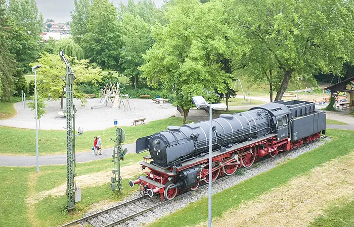 Luftaufnahme der Treuchtlinger Denkmalslok inklusive dahinter liegendem Spielplatz mit Besuchern inmitten von Grün.