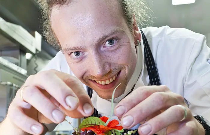 Ein junger Koch mit schwarzer Schürze und weißem Hemd lächelt in die Kamera. er dekoriert ein Tatar mit grünem Salat und einer Blume.