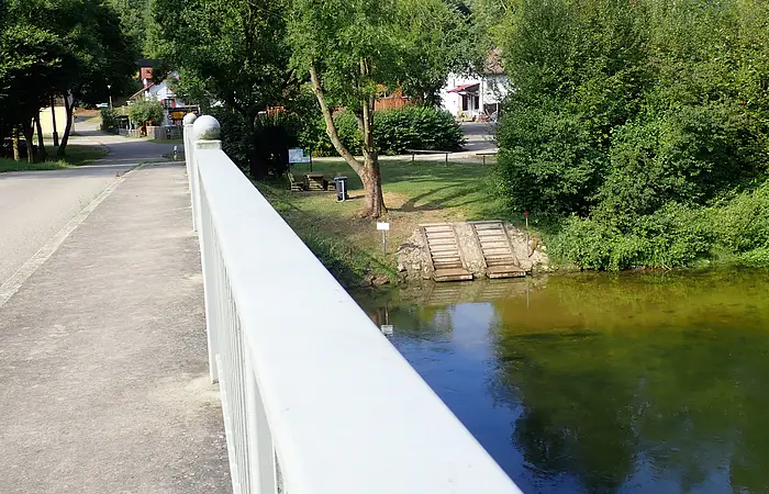 Brücke mit weißem Geländer über Fluss, links Straße, rechts Bäume und Häuser im Hintergrund