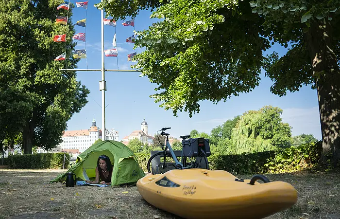 Ein Kajak und ein grünes Zelt sind auf dem Bootsübernachtungsplatz. Im Hintergrund grüne Bäume, ein Fahrrad und hellblauer Himmel.