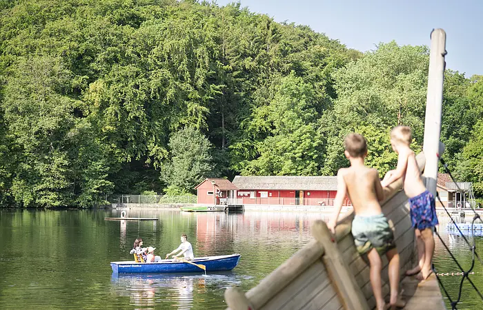 Zwei Kinder stehen auf dem Piratenschiff und beobachten ein Ruderboot, dass auf dem Waldsee in Wemding fährt.