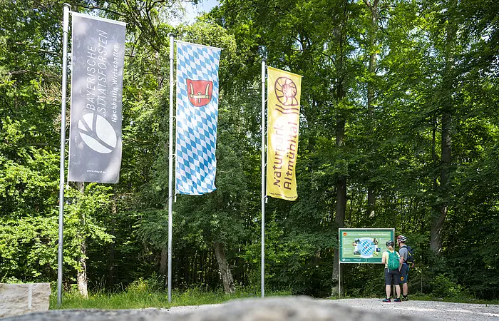 Zwei Wanderer stehen an der Infotafel am Geographischen Mittelpunkt Bayerns bei Kipfenberg und lesen interessiert. Links von ihnen sind drei Fahnen gehisst.