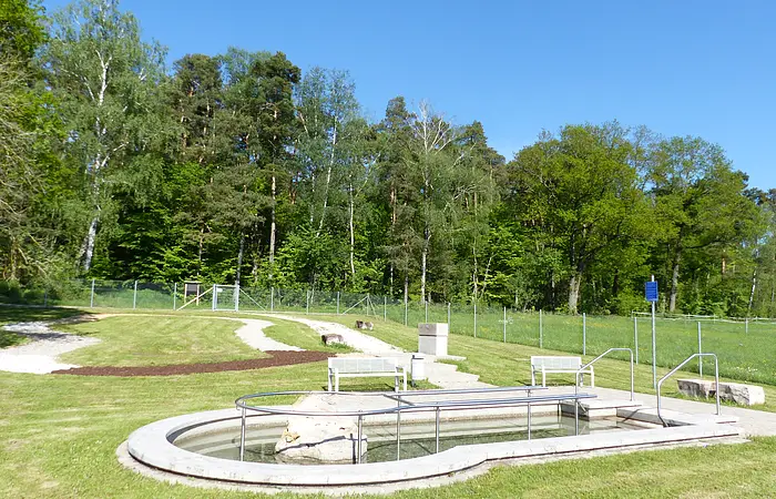 Rundes Kneippbecken in Monheim mit einem Geländer und einem Barfußpfad auf einer Wiese, umgeben von einer großen Grünfläche und Bäumen und Sitzmöglichkeiten. Ein Zaun im Hintergrund.