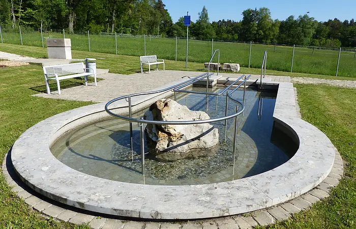 Rundes Kneippbecken in Monheim mit einem Geländer und einem Barfußpfad, in der Mitte ein großer weißer Stein. Umgeben von einer Wiese, darauf Sitzmöglichkeiten und eine große Grünfläche mit Bäumen hinterhalb, abgetrennt mit einem Zaun.
