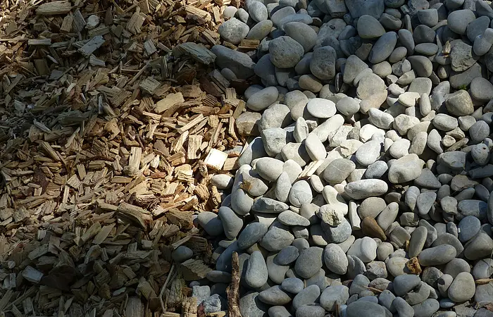 Holzhackschnitzel links neben einer Fläche mit kleinen, grauen Steinen von der Sonne bestrahlt.