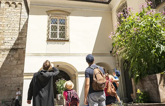 Eine Familie steht mit einem Mönch vor dem Eingang zum Kloster Plankstetten. Der Mönch erklärt der Familie etwas von der Geschichte des Gebäudes. Er hat seinen Arm gehoben und zeigt auf etwas.